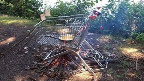 Einkaufswagen dient als Grill über offenem Feuer im Freien, Bratpfanne darin, umgeben von Bäumen und Gras.