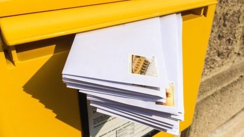 Ein gelber Briefkasten hat einige versendete Briefe mit Marken, die herausragen, während der Kontext eine sonnige Umgebung ist.