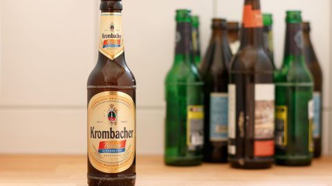 Eine Flasche alkoholfreies Bier steht auf einem Holztisch, umgeben von leeren, grünen und braunen Bierflaschen.
