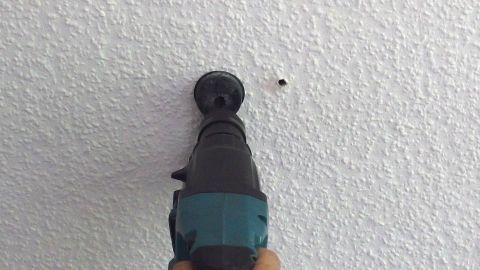 Ein Bohrer durchdringt eine raue Wand, während eine Person ihn hält; der Kontext ist ein normaler Innenraum.