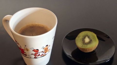 Kaffee und Kiwi: Innerhalb von 10 Minuten sind die Kopfschmerzen und die Übelkeit verschwunden. Eine Tasse Kaffee steht neben einer Kiwi auf einem schwarzen Teller, auf einem glatten Tisch, ideal für Kater und Kopfschmerzen.