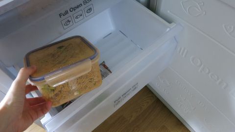Eine Hand hält eine transparente Box mit Essen, während sie in ein offenes Kühlschrankfach legt, um Lebensmittel frisch zu halten.