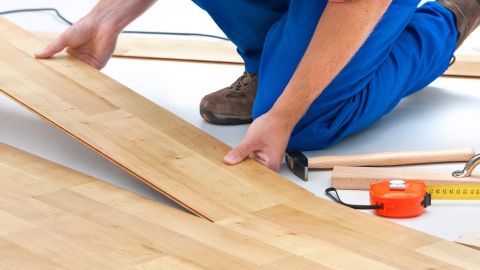 Person installiert Laminat, kniet auf Boden; Umgebung: Werkzeug wie Maßband, Hammer und Holzbrett.