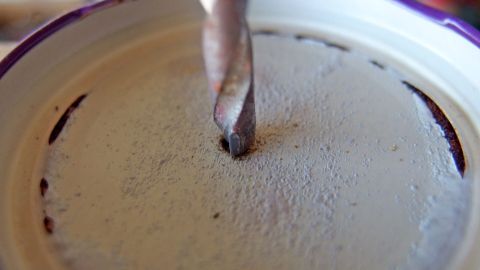 Beim Über-Kopf-Bohren hilft ein einfacher Trick: Fange den Bohrstaub mit einem umgedrehten Deckel, wie hier gezeigt, auf. So bleibt der Boden sauber und du kannst effizienter arbeiten. Ein Bohrer durchdringt langsam einen Deckel, während Metallspäne um die Bohrstelle herum verstreut sind.