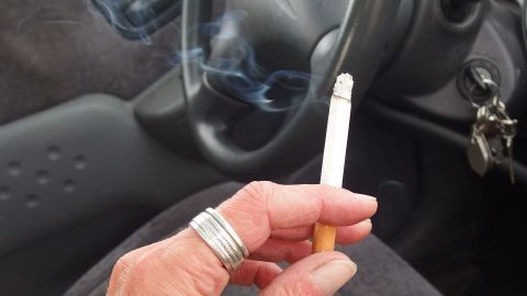 Eine Hand hält eine Zigarette, während Rauch im Auto aufsteigt, was den typischen Rauchgeruch im Innenraum verstärken kann.