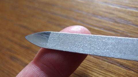 Ein Finger mit Sekundenkleber ist in Kontakt mit einer Nagelfeile, die ihn vorsichtig bearbeitet, auf einem Holztisch.