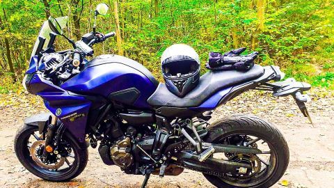 Ein Motorrad steht auf einem Waldweg, während ein Helm und Handschuhe auf dem Sitz liegen. Die Umgebung ist grün und herbstlich.