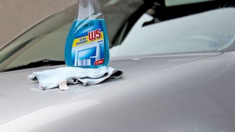 Eine Flasche Glasreiniger steht auf der Motorhaube eines Autos, während ein sauberes Tuch daneben liegt, bereit zum Putzen.