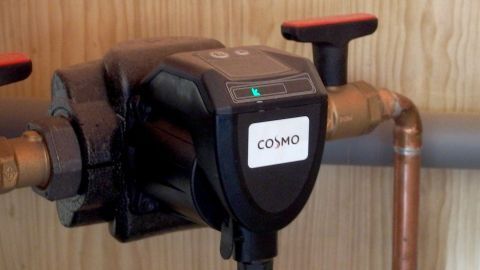 Eine Heizungsumwälzpumpe mit der Aufschrift "COSMO" ist an einem Kupferrohr installiert, umgeben von Holzverkleidung.