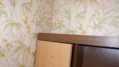 Mit einem Glasreiniger lassen sich Tapeten am besten lösen. Ein Holzregal steht unter einer Tapete mit floralen Mustern. Der Raum wirkt hell und freundlich.