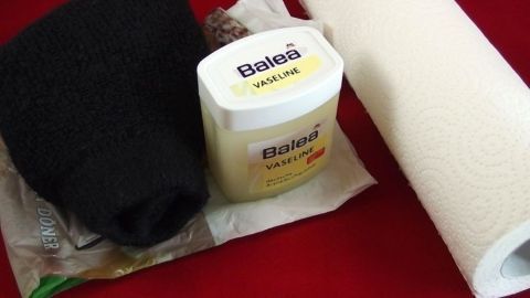 Ein schwarzes Peeling-Handschuh liegt neben einer Creme für Hornhautpflege und einem Papiertuch, alles auf rotem Hintergrund.