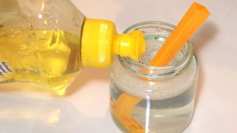Rasierklingen bleiben länger scharf wenn man sie nach Gebrauch in einem Becher mit Wasser und Spülmittel aufbewahrt. Ein gelbes Flaschenbehälter gießt eine Flüssigkeit über eine orange Rassierklinge in einem Glasbehälter mit Wasser.