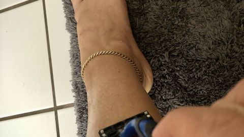 Eine Hand hält einen Rasierer, der über das Bein gleitet, während sich das Fuß und ein Teppich im Hintergrund befinden.