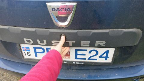 Ein Finger berührt die Heckklappe eines blauen Dacia Duster, während das Auto auf einem Parkplatz steht.