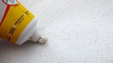 Mit „Rauhfaser Reparatur Moltofill“ lassen sich Bohrlöcher und gerissene Fasern auf der Rauhfasertapete ohne Probleme ausbessern. Eine Tube mit Wandfarbe liegt auf einer Rauhfasertapete und ist bereit, kleine Schäden auszubessern.