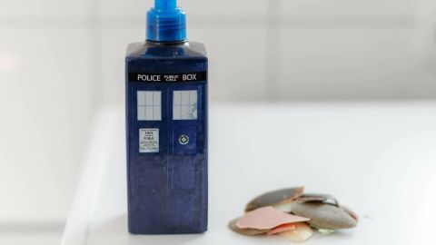 So verwertet man mithilfe eines Flüssigseifen-Pumpenspenders und Wasser, herumliegende alte Seifenreste. Ein Seifenspender in Form einer blauen Telefonzelle steht neben bunten Seifenresten auf einer weißen Oberfläche.