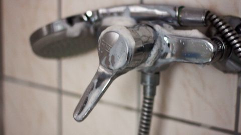 Kalk mit Klarspüler entfernen: Füllt man den Klarspüler in eine leere Glasreiniger Sprühflasche um, kommt man auch an unzugängliche Stellen (Dusche, Wasserhähne etc.). Ein glanzender Wasserhahn wird in einem Badezimmer betrachtet, um Kalk mit Klarspüler zu entfernen. Fliesen umgeben den Hahn.