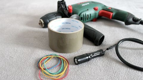 Eine Bohrmaschine liegt zusammen mit Klebeband, einem Taschenlampen und Gummibändern auf einem hellen Untergrund.