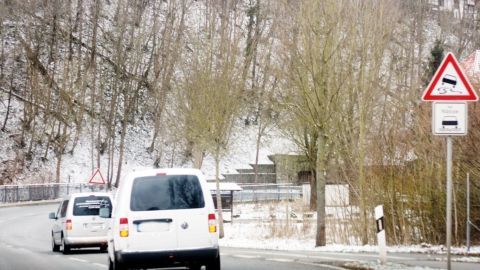 Zwei Fahrzeuge fahren auf einer schneebedeckten Straße, während ein Warnschild auf winterliche Bedingungen hinweist.