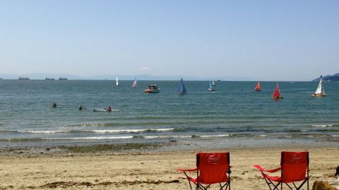Rote Stühle stehen am Strand, während Boote mit bunten Segeln im ruhigen Wasser vorbeifahren. Ein entspannender Urlaub.