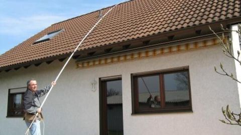 Ein Mann entfernt Moose und Flechten von einem Dach mit einem langen Besenstiel, während ein Haus mit Fenstern im Hintergrund sichtbar ist.