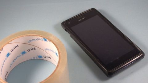 Ein Handy liegt auf einem hellblauen Untergrund, während eine Rolle Klebefolie danebensteht, bereit zum Basteln einer Schutzfolie.
