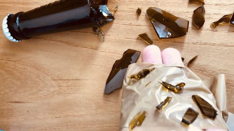 Ein zerbrochener Glasflasche liegt auf einem Holzuntergrund, während jemand mit einem Handschuh die Glassplitter aufnimmt.
