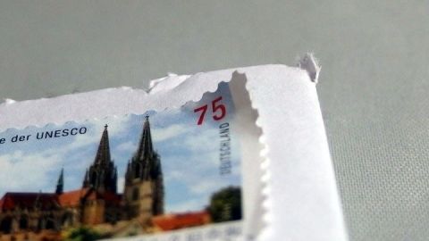 Eine Briefmarke wird vorsichtig von einem Kuvert gelöst; sie zeigt eine Kirche, Text: "Kulturdenkmal der UNESCO", "75", "DEUTSCHLAND".