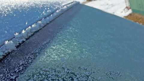 Eine gefrorene Autoscheibe wird von lauwarmem Wasser bearbeitet, um das Eis zu entfernen, während Schnee im Hintergrund liegt.