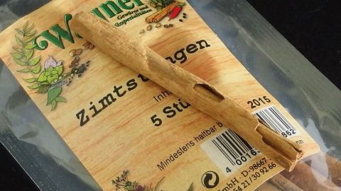 Zimtstangen liegen in einer transparenten Verpackung, umgeben von Kräuterillustrationen und einem Holzdesignhintergrund.