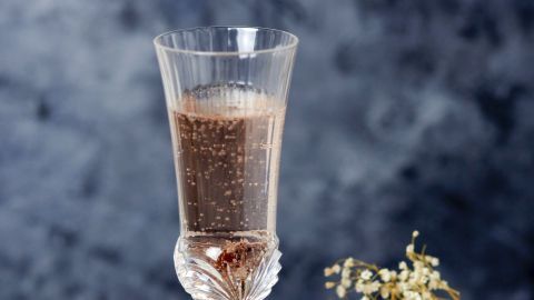 Ein stilvolles Glas Schaumwein sprudelt und steht auf einem Holzbrett, umgeben von dekorativen Blüten und Rosinen.