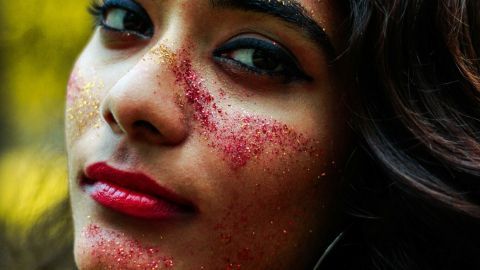 Eine Frau mit glitzerndem Makeup auf ihrem Gesicht schaut in die Kamera und steht in einer natürlichen Umgebung.