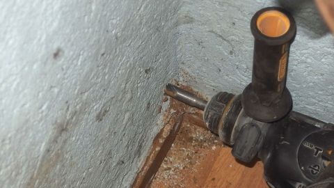 Ein Bohrer wird verwendet, um ein zu großes Dübelloch in der Wand zu bearbeiten. Die Umgebung zeigt Holz und eine Wand.