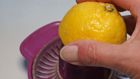 Augenringe? Kein Problem, einfach eine Zitrone auspressen und eine geringe Menge auf die betroffenen Stellen einmassieren, aber ACHTUNG, darf nicht ins Auge gelangen, sonst brennt es höllisch. Eine Zitrone wird mit einer Hand auf einem Entsafter gedrückt, um Saft zu gewinnen, während sie auf einem Tisch liegt.