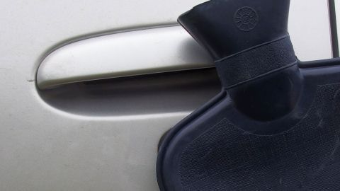 Eine Wärmflasche liegt an einem gefrorenen Autotürschloss, um es aufzutauen. Im Hintergrund ist das silberne Auto sichtbar.