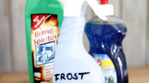 Eine Sprühflasche mit der Aufschrift "FROST SCHUTZ" steht vor weiteren Reinigungsflaschen auf einem Holztisch.