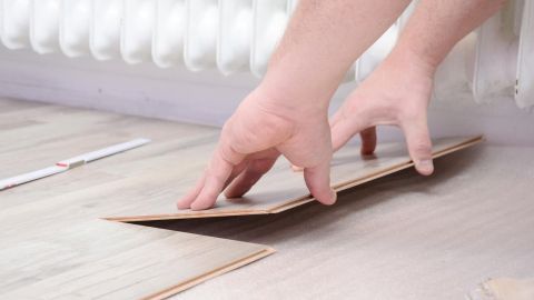 Hände legen ein Klicklaminat-Element auf den Boden, während im Hintergrund ein Heizkörper zu sehen ist.