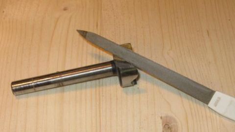 Ein Forstnerbohrer liegt auf einer Holzoberfläche, während eine Nagelfeile daneben zum Schärfen verwendet wird.