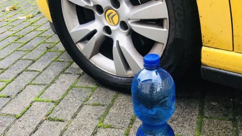 Eine blaue PET-Flasche steht am Bordstein neben einem gelben Auto, das auf einem gepflasterten Weg parkt.