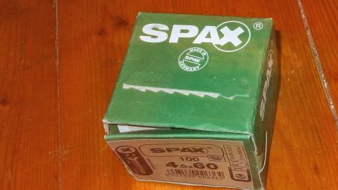 Ein grüner Karton mit der Aufschrift „SPAX“ steht auf einem knarrenden Fußboden aus Holz, umgeben von natürlichem Licht.
