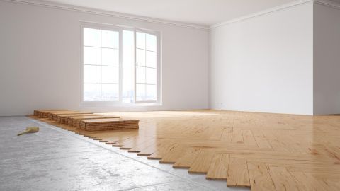 Holzparkett wird in einem leeren, hellen Raum am Fenster verlegt; ein Stapel Planken und Hammer liegen auf dem Betonboden.