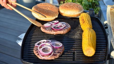 Saftige Burgerpatties grillen auf dem Balkon, während frische Brötchen und Maiskolben bereitliegen.