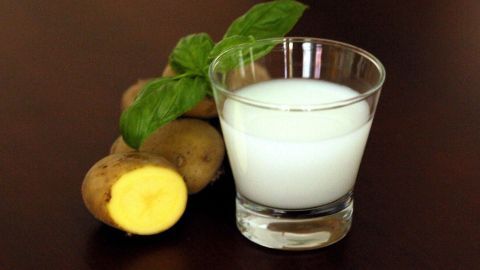 Ein Glas mit klarem Kartoffelsaft steht neben frischen Kartoffeln und einem grünen Blatt im Hintergrund.
