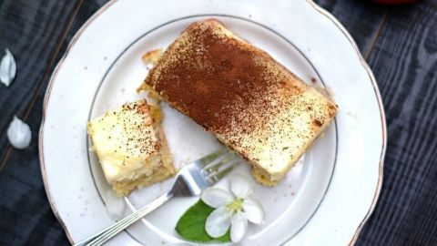 Apfeltiramisu auf einem Teller präsentiert sich neben frischen Äpfeln und weißen Blüten auf einem dunklen Holztisch.