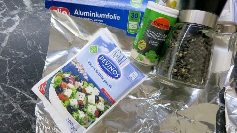 Auf einer Tischoberfläche liegen Aluminiumfolie, ein Gewürzmühlenbehälter und eine Verpackung für Feta-Käse, bereit zum Kochen.