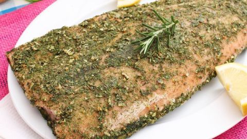 Ein Lachs im Kräutermantel liegt auf einem weißen Teller, garniert mit Zitronenscheiben und Kräutern, auf einem bunten Tisch.