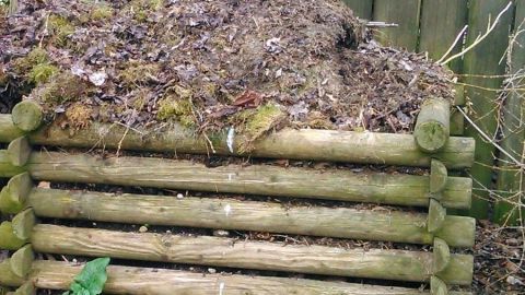 Ein Holzkomposter steht im Garten mit einem Haufen organischen Materials darauf, umgeben von einem Lattenzaun.