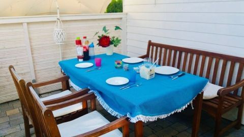 Holzmöbel stehen auf einer Terrasse, gedeckt mit blauem Tischtuch und Geschirr, umgeben von weißem Sichtschutz und Sonnenschirm.