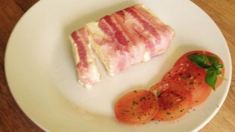 Ein Stück Bacon-Schafskäse liegt auf einem weißen Teller, begleitet von frischen Tomatenscheiben und Basilikum.