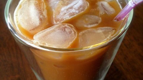 Ein Glas Eiskaffee mit Eiswürfeln steht auf einem Holztisch. Ein Strohhalm steckt darin, bereit zum Trinken.